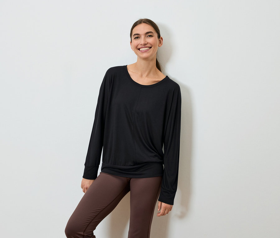 Une femme souriante portant un haut noir à manches longues et des leggings marron pose contre un mur.