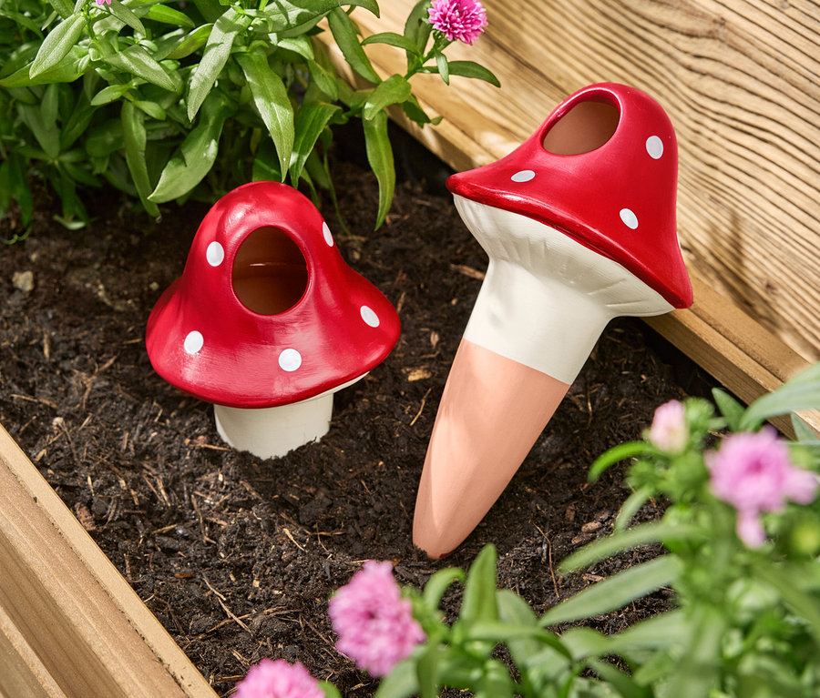 Deux boules d'arrosage rouges en forme de champignons avec des points blancs se trouvent dans une jardinière entre des fleurs et des plantes.
