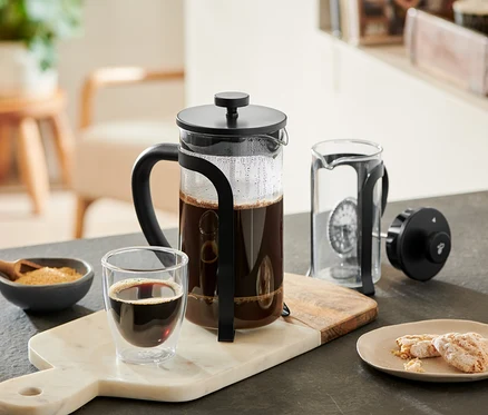 Une cafetière à piston 800 ml, 6 tasses, noire, remplie de café, est posée sur une planche en marbre à côté d'un verre de café, d'un bol de sucre et de biscuits. Une autre cafetière à piston est démontée à côté.
