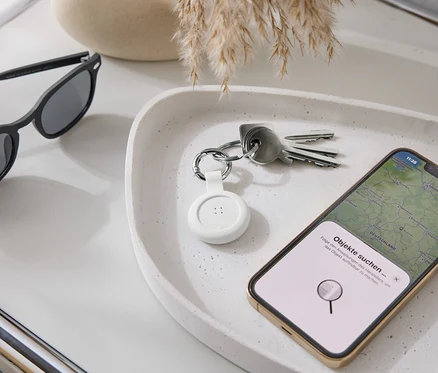 Sur un plateau reposent des lunettes de soleil, des clés, un traceur blanc et un smartphone avec une application de carte.