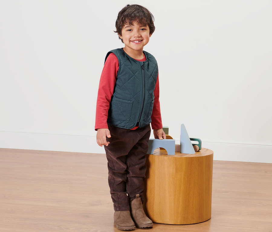 Un garçon se tient à côté d'une table en bois et porte un gilet matelassé réversible pour enfant.