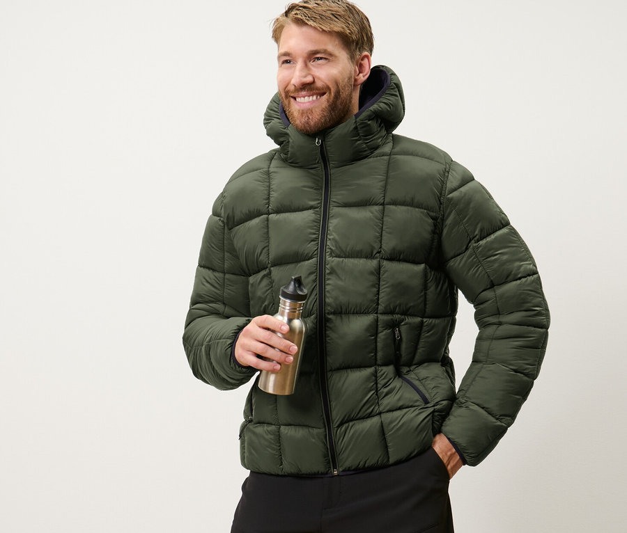 Un homme tient une bouteille d'eau et porte une veste matelassée kaki.