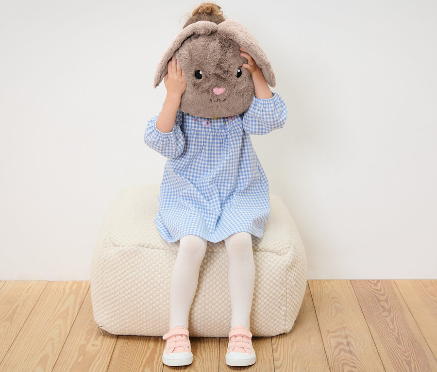 Fillette assise sur un pouf tenant un coussin en peluche petit lapin devant son visage.