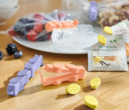 Sur une table en bois, il y a 15 clips arôme de différentes couleurs, des sachets de fruits, de noix et de sucre vanillé.