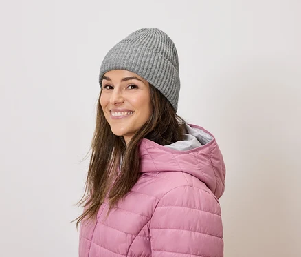 Une femme aux cheveux bruns sourit et porte un bonnet en tricot gris et une doudoune rose.