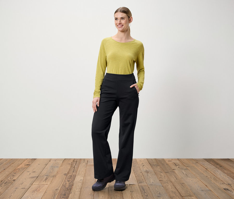 Une femme se tient sur un plancher en bois. Elle porte un chemisier vert à manches longues et un pantalon noir.