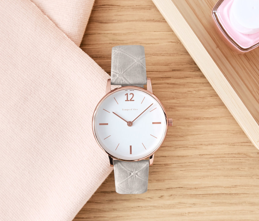 Une montre-bracelet avec bracelet en cuir est posée sur une table en bois à côté d'un tissu rose et de cosmétiques.
