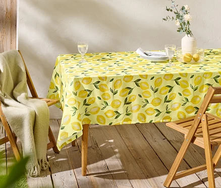 Une table à manger avec une nappe jaune à motif de citrons, deux verres, des assiettes et deux chaises en bois.