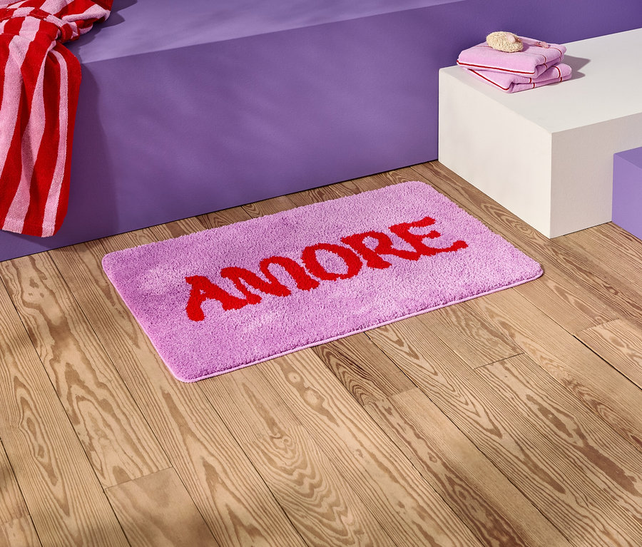 Sur un plancher en bois se trouve un tapis de bain lilas avec l'inscription rouge "AMORE".