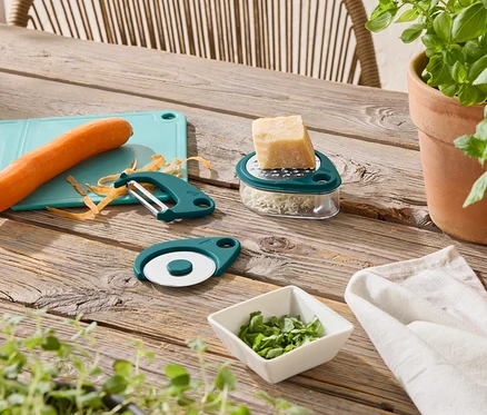 Sur une table en bois se trouvent une planche à découper bleue avec une carotte, un éplucheur vert, un coupe-pizza vert, une râpe à fromage avec du fromage et un bol d'herbes.
