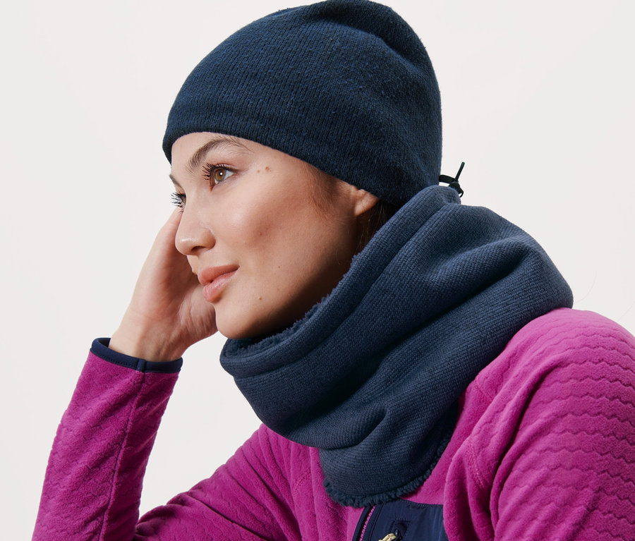 Une femme appuyée sur un banc en bois porte une écharpe-tube en maille polaire bleue et un bonnet bleu.