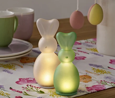 Deux lapins LED sont posés sur un chemin de table à fleurs, avec des tasses et des œufs de Pâques en arrière-plan.