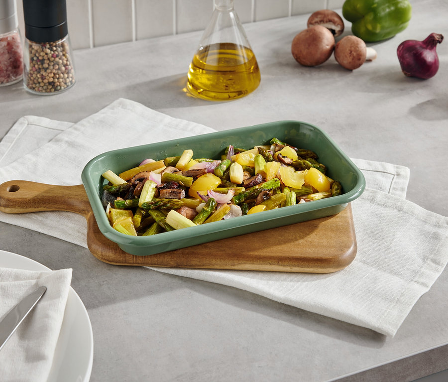 Plat de cuisson vert avec des légumes cuits sur une planche à découper avec une serviette. À l'arrière-plan, on trouve des champignons, des poivrons, des oignons et des épices.