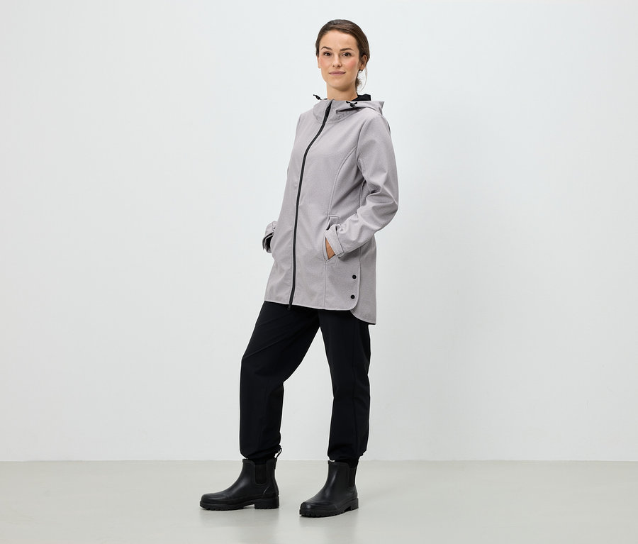 Une femme se tient debout, les mains dans les poches d'une veste à capuche grise, un pantalon noir et des bottes noires.