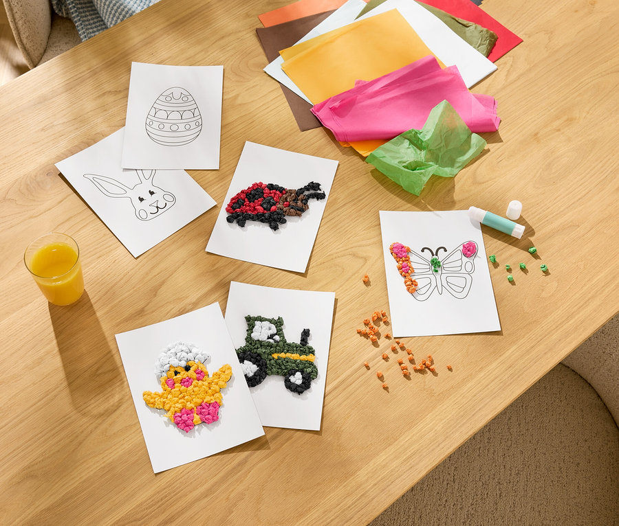 Sur une table en bois se trouvent des travaux manuels, notamment des cartes collées de poussins, de tracteurs, de papillons et de tortues. Du papier de soie coloré, un bâton de colle et un verre de jus sont également visibles.