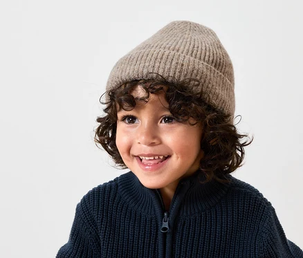 Garçon souriant avec des cheveux bouclés bruns portant un bonnet en laine beige et un cardigan zippé en tricot bleu marine.