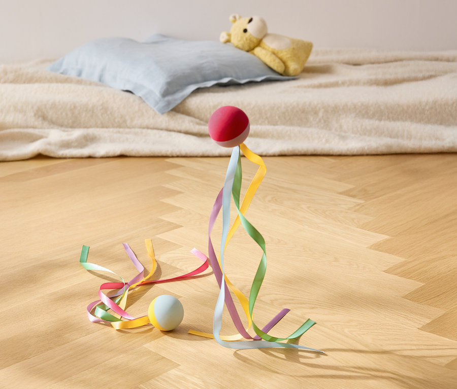 Une balle rouge repose avec des rubans colorés sur un plancher en bois, avec un oreiller et un ours en peluche en arrière-plan.