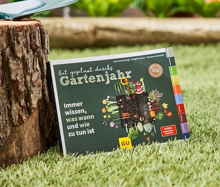 Le livre « Gut geplant durchs Gartenjahr » est posé sur l'herbe à côté d'une souche d'arbre sur laquelle repose une pelle de jardin.