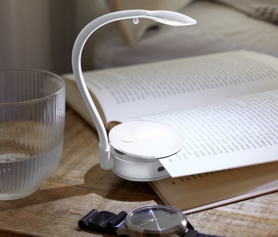 Sur une table de chevet reposent un livre ouvert éclairé par une lampe de lecture LED, un verre d'eau et une montre.