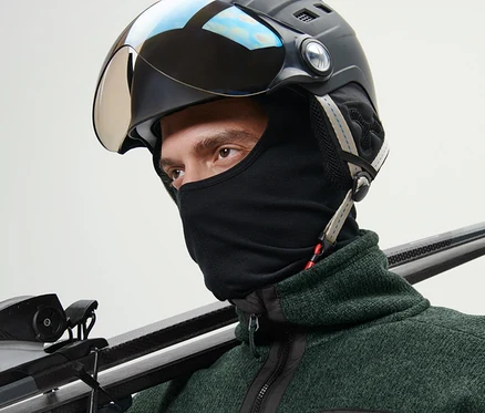 Un homme portant un casque de ski et une cagoule de performance tient des skis sur son épaule.