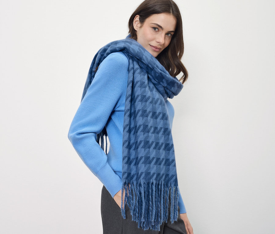 Une femme aux cheveux bruns porte un pull bleu et une écharpe bleue à carreaux.