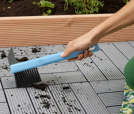 Une femme nettoie des dalles de terrasse grises avec une brosse bleue.