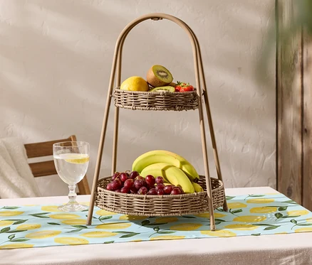 Un panier à fruits à deux étages en osier est posé sur une table avec une nappe à motif de citron. Le panier supérieur contient un citron, un kiwi et des fraises, tandis que le panier inférieur contient des bananes et des raisins rouges.