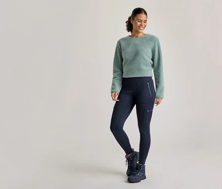Une femme souriante portant un pull duveteux vert clair, un legging cargo bleu foncé et des chaussures de randonnée bleues foncées se tient debout, une jambe pliée.