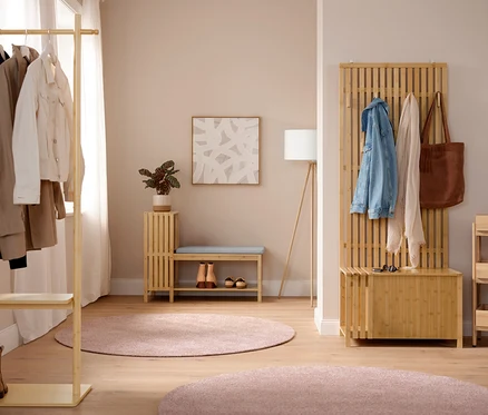 Un hall d'entrée lumineux avec un porte-manteau avec tablette où pendent des vestes. Au centre, un banc avec compartiment à chaussures et étagères en finition lamellé avec un coussin et des chaussures en dessous, à côté d'un lampadaire trépied en bois. À droite, une garde-robe avec banc, aspect lamellé, avec une veste en jean, une écharpe et un sac. À côté, une étagère sur pieds avec 3 compartiments. Deux tapis ronds roses sont posés au sol.