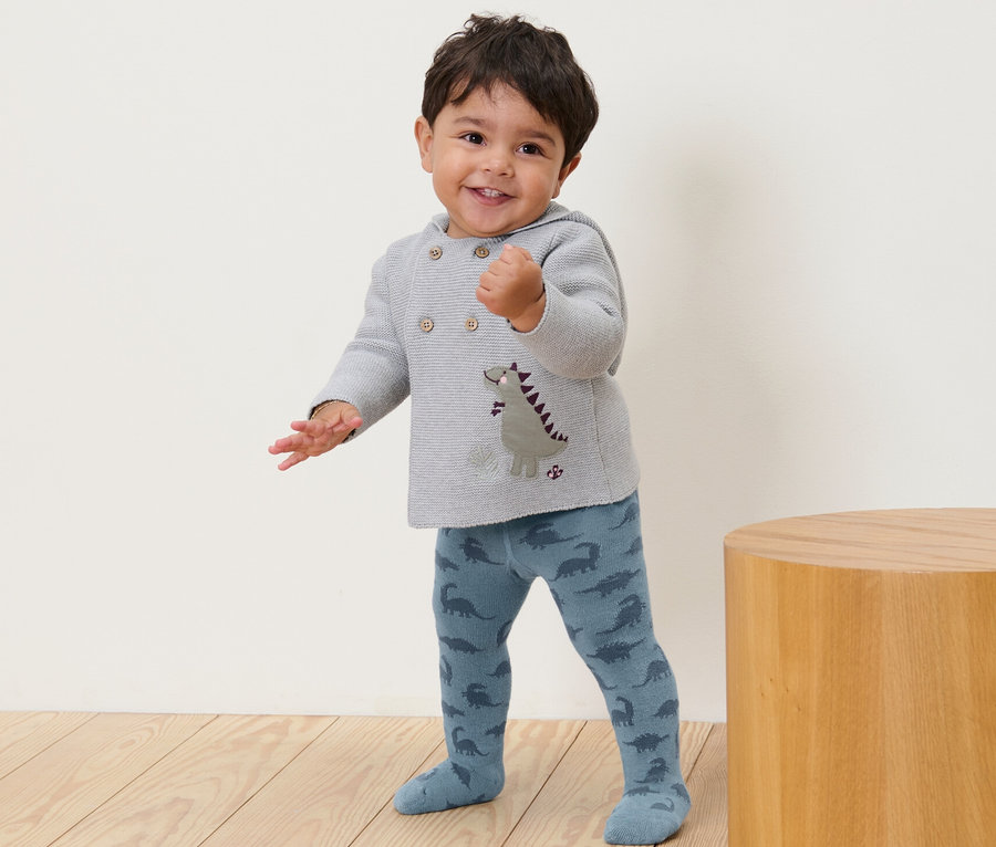 Un jeune enfant souriant se tient debout et porte des collants bleus à motifs de dinosaures.