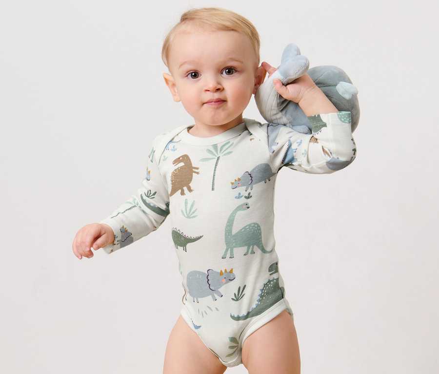 Un bébé tient une peluche et porte un 3 bodys manches longues, dinosaure.