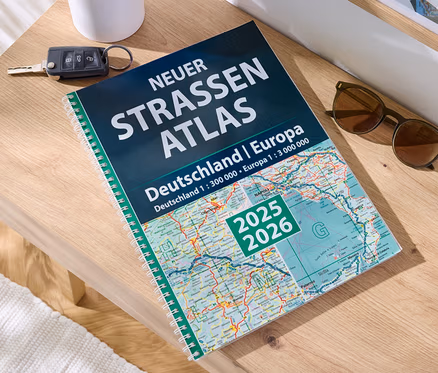 Sur une table en bois sont posés un livre 'Livre : nouvel atlas routier Allemagne/Europe 2025/2026', une clé de voiture et des lunettes de soleil.