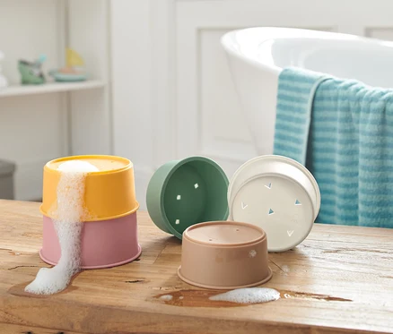 Sur une table en bois, des verres empilables pour jouer dans le bain, dont deux sont empilés avec de la mousse qui s'échappe du verre supérieur.