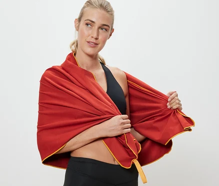 Une femme blonde drape une serviette de bain d’extérieur orange d'environ 100 x 180 cm autour de ses épaules.