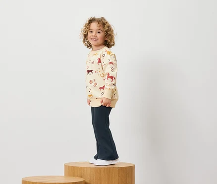 Garçon aux cheveux bouclés portant un sweat-shirt à imprimé cheval et un pantalon bleu marine, debout sur un socle en bois.