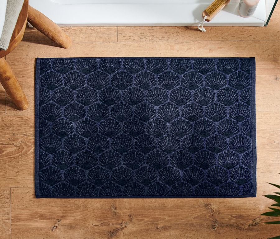 Tapis de douche bleu marine « Coquillage » posé sur un plancher en bois à côté d'un tabouret et d'une plante.