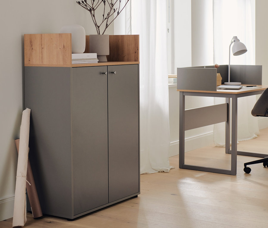 Un secrétaire gris avec bord latéral surélevé et un buffet haut à 2 portes se trouvent l'un à côté de l'autre. Une lampe de table en métal est posée sur le secrétaire.