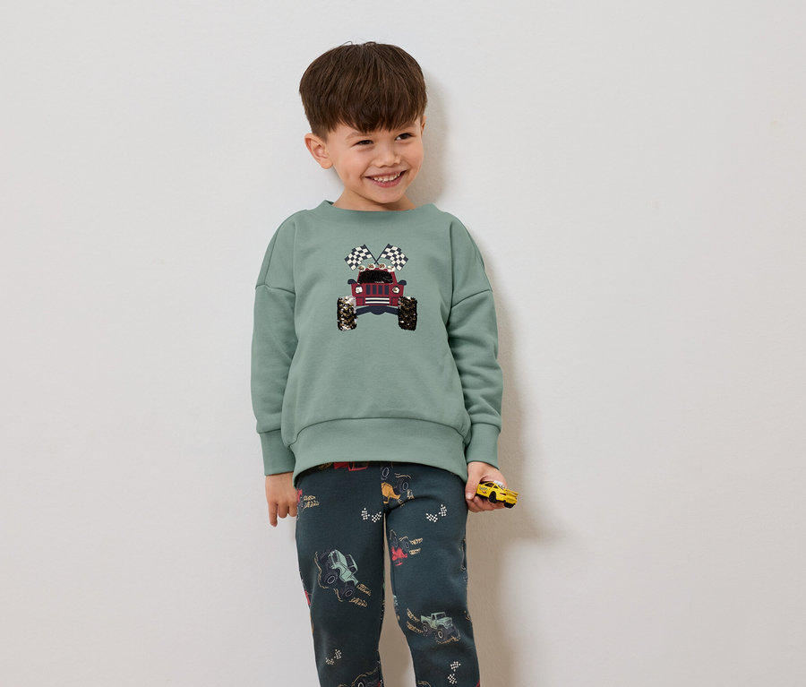 Un garçon porte un pull vert avec un imprimé de jeep et de drapeaux à damier, ainsi qu'un pantalon foncé avec un motif de jeeps et tient une voiture jaune.