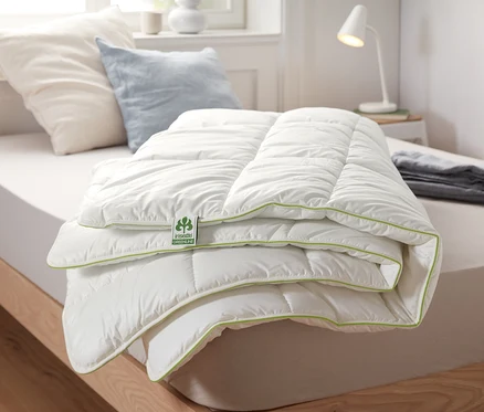 Une couette thermique irisette® greenline pliée avec une surface blanche et une bordure verte est posée sur un lit.