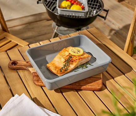 Un morceau de saumon est posé dans un plat à gratin gris sur une table en bois, à côté d'un barbecue.