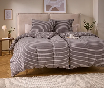 Un lit avec une parure de lit en molleton gris et blanc à rayures, avec un motif quadrillé au verso. Deux oreillers assortis au motif rayé. Une tasse blanche est posée sur la couette. À gauche, une table de chevet en bois avec une lampe et un vase, et un tapis clair au sol.