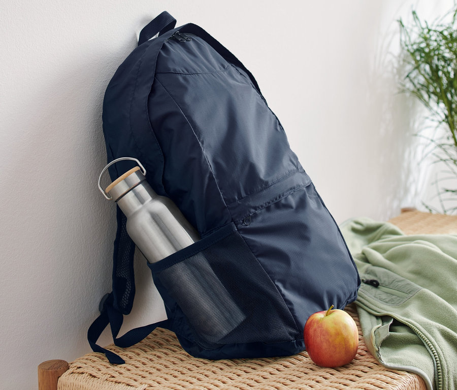 Un sac à dos pliable bleu marine appuyé contre un mur blanc sur un banc en osier. Une bouteille d'eau argentée est dans la poche latérale du sac à dos, et une pomme est posée à côté du sac.