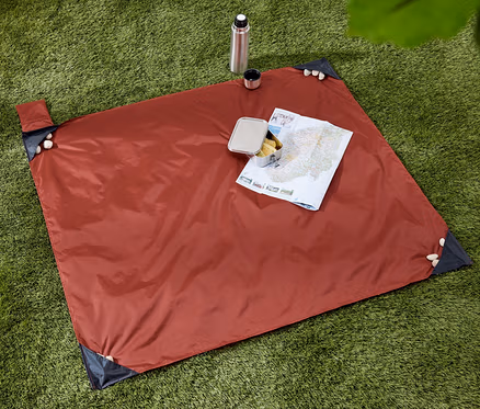Sur l'herbe verte se trouve une mini-couverture de pique-nique avec une carte et une collation. Une bouteille thermos et une tasse sont à côté.