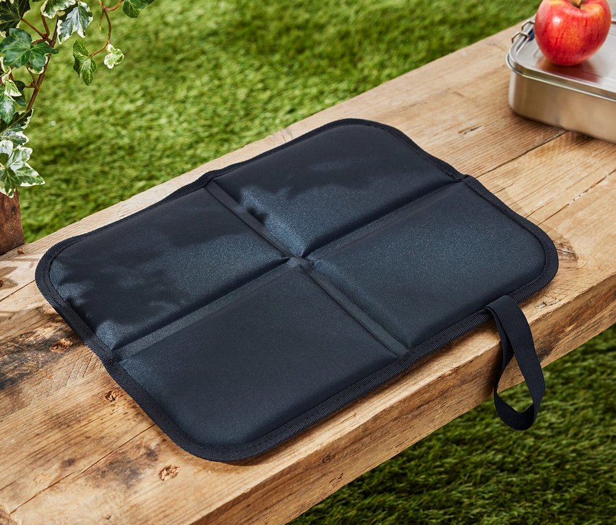 Un coussin d’assise thermique pliable est posé sur un banc en bois à côté d’une pomme et d’une boîte à lunch.