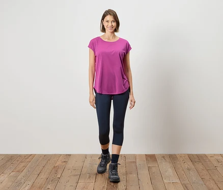 Une femme se tient sur un plancher en bois devant un mur blanc. Elle porte un t-shirt rose, des leggings bleus et des bottes grises.