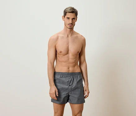 Un homme torse nu porte un caleçon boxer gris et se tient devant un fond clair.
