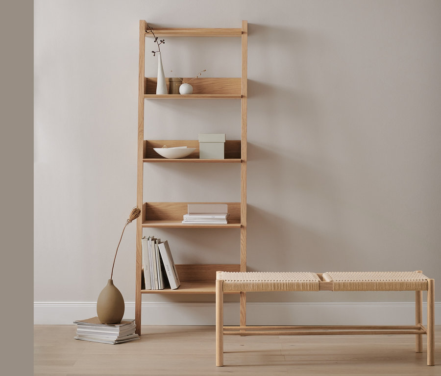 Un rayonnage échelle en chêne avec des décorations et des livres à côté d'un banc avec matière tressée.