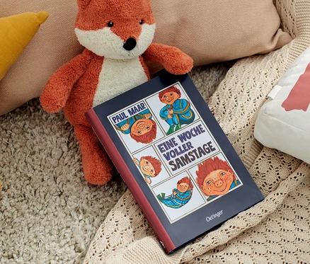 Un renard en peluche est posé à côté du livre « Das Sams – une semaine pleine de samedi » et de coussins.