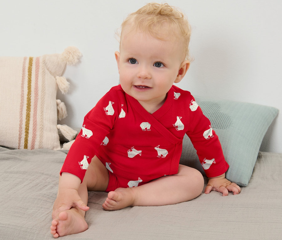 Une petite fille blonde porte un body rouge à manches longues avec un motif d'ours polaire et est assise sur une couverture grise.