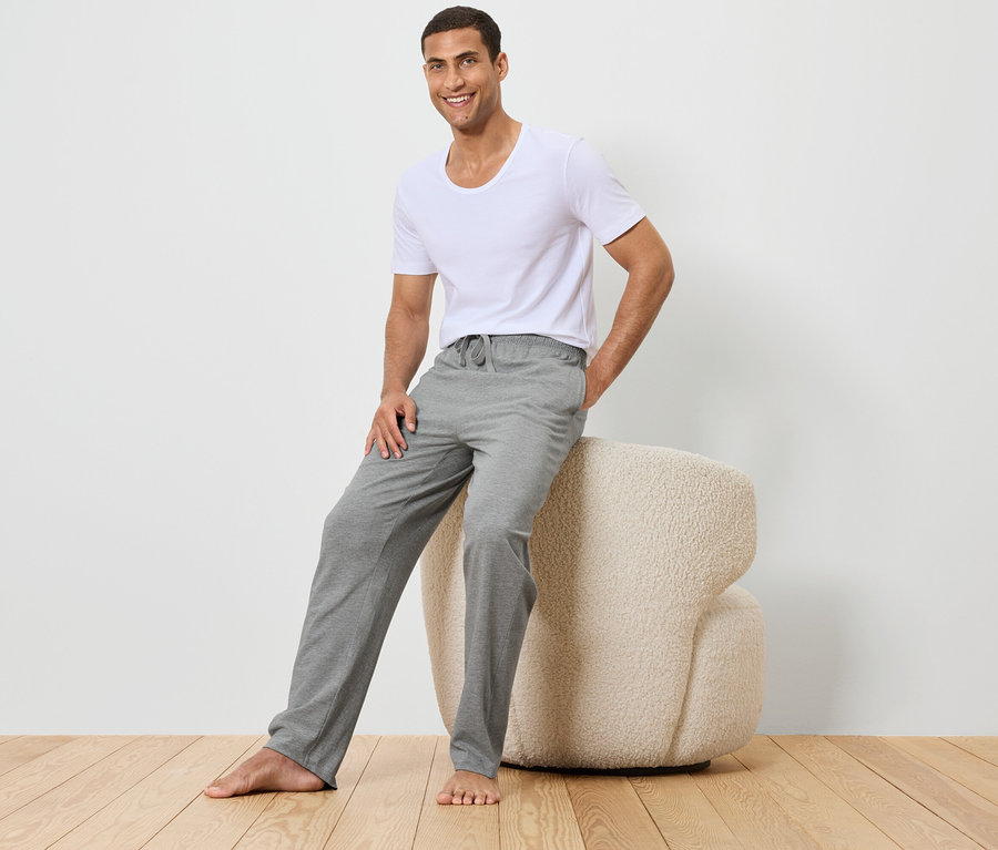 Un homme est assis sur un tabouret beige, portant un t-shirt blanc et un pantalon gris.